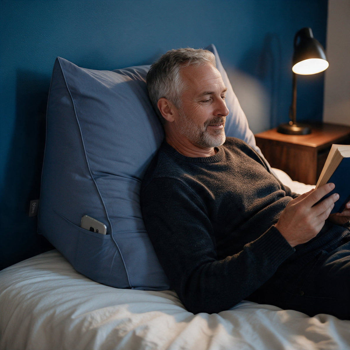 Man leest 's avonds tegen het leeskussen Bline, kleur Blauw, met telefoon in het zijvak